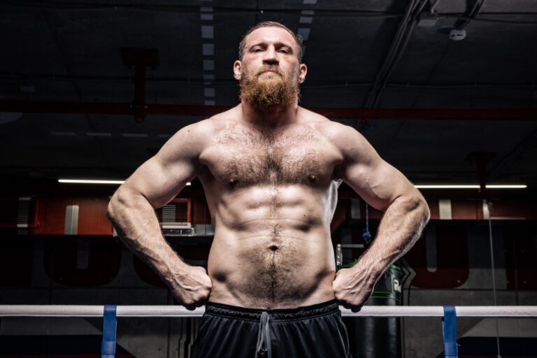 Strong shirtless boxer with a beard stands in a gym ring, showcasing power and fitness.