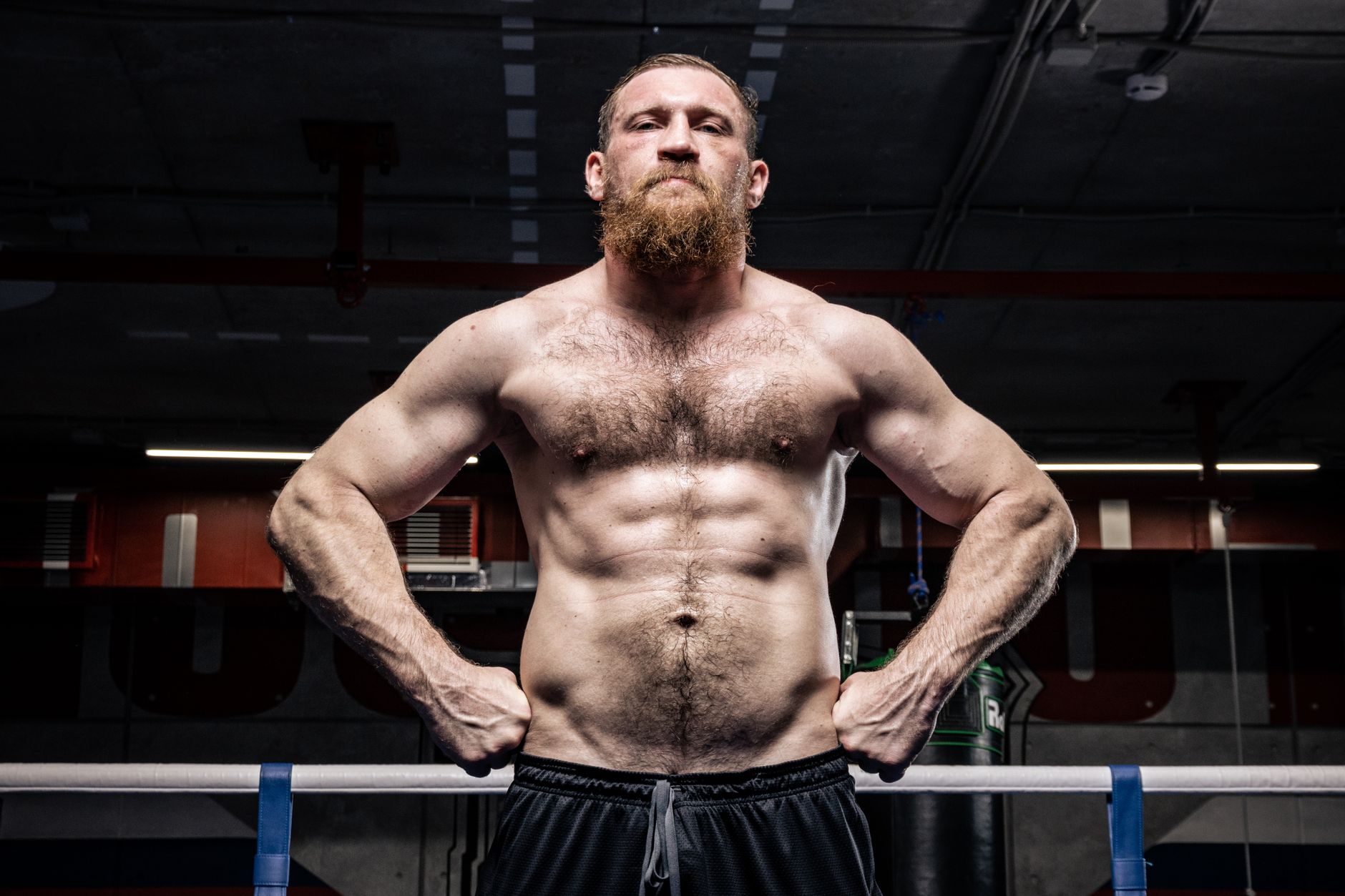 Strong shirtless boxer with a beard stands in a gym ring, showcasing power and fitness.