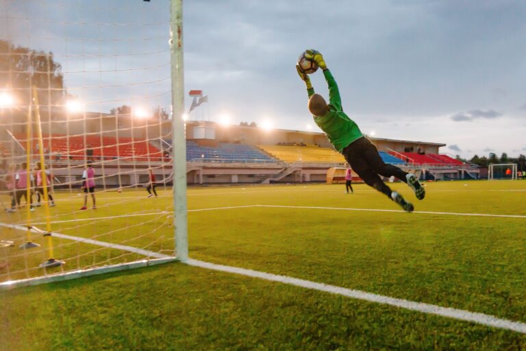 Goalkeeper making a diving save on a soccer field during a match at sunset.