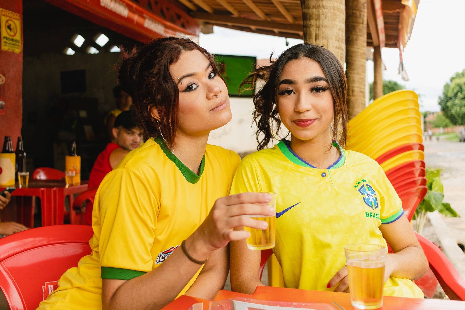 Two women in Brazil jerseys enjoying drinks at an outdoor venue.