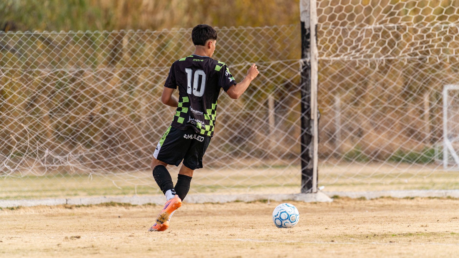 A teenager in sports uniform shooting a soccer ball towards a goal on an outdoor field.