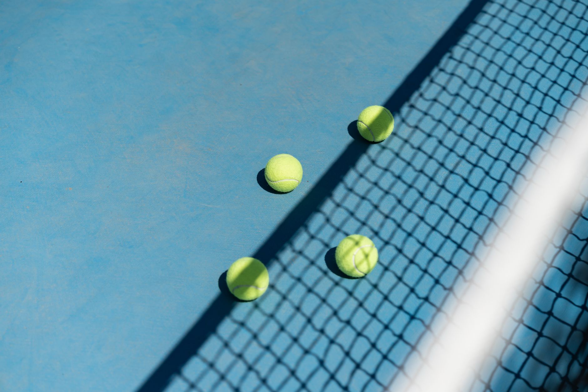 Three tennis balls scattered on a blue court with a shadowed net.
