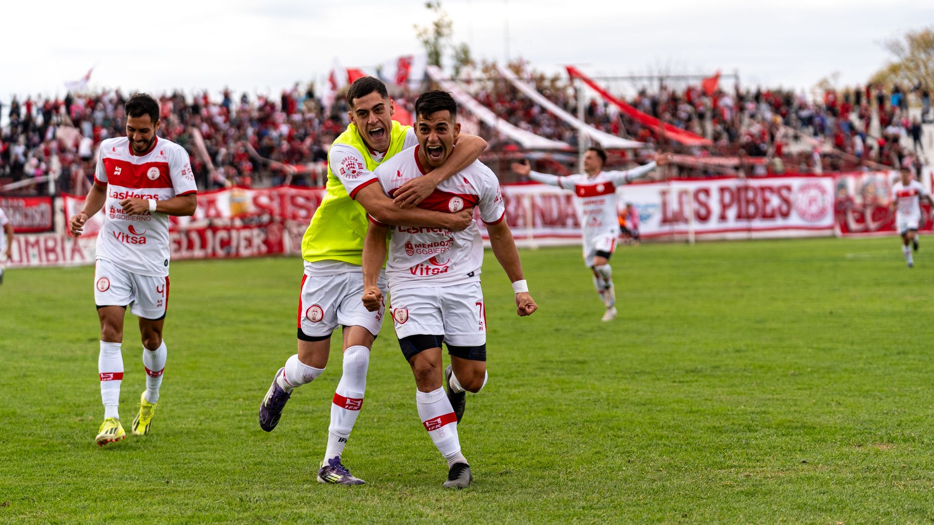 Soccer players celebrating a goal on an outdoor field with cheering crowd.