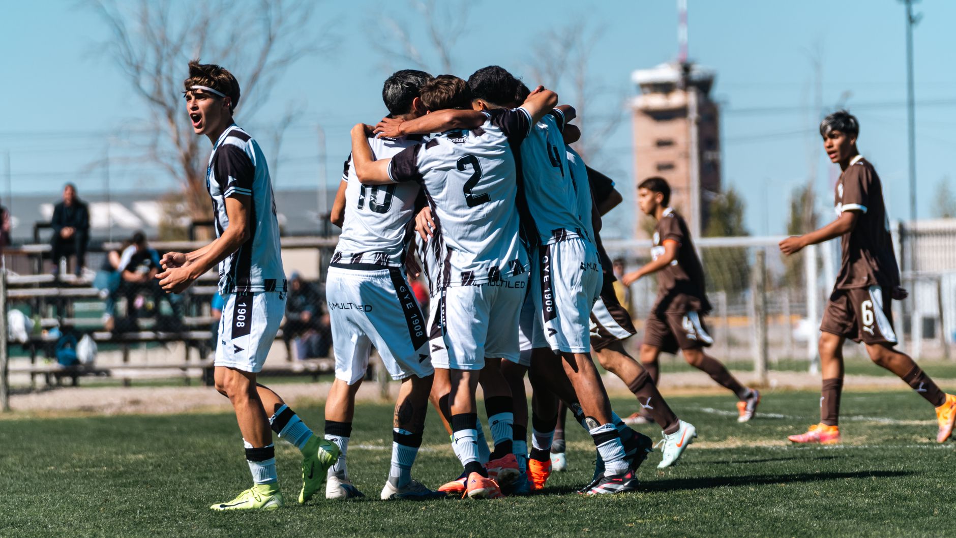 Teenage soccer team celebrates a goal on a sunny day during an outdoor match on a grassy field.